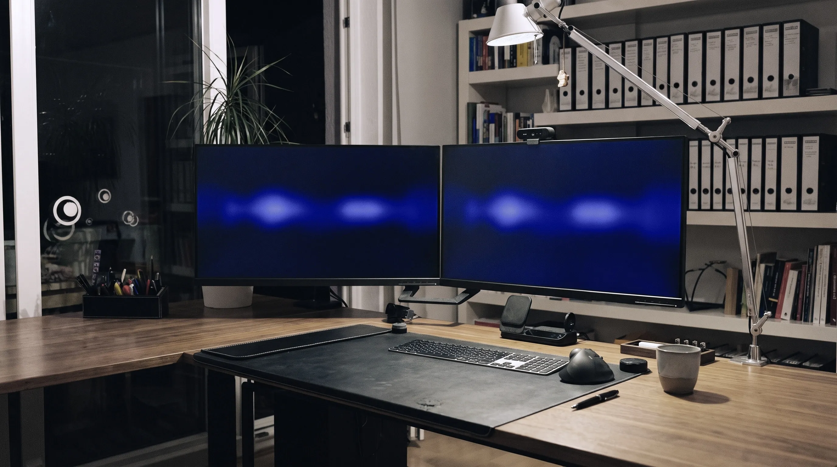 The desk in the evening: two screens above a solid wood top, next to them the shelf of box files and a plant at the dark window
